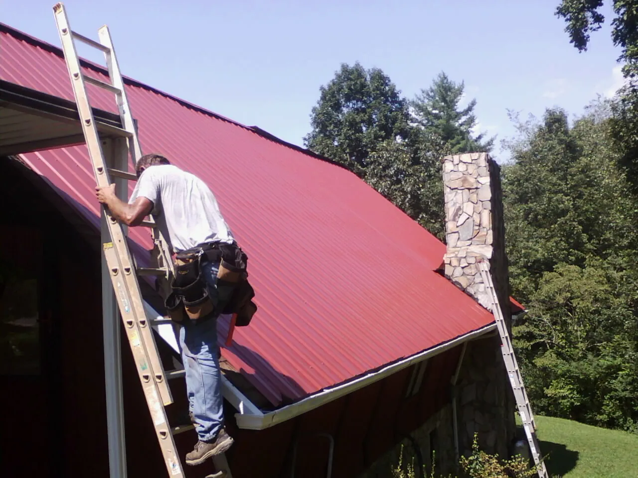 Roofer on a ladder beside a red metal roof and stone chimney — Soffit & Fascia Repair by Roofing Brothers in Kodak